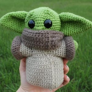May include: A handmade crochet doll of a character with large ears and black eyes. The doll is primarily green, with a brown and beige outfit. The doll is held against a backdrop of green grass.