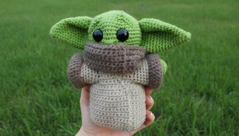 May include: A handmade crochet doll of a character with large ears and black eyes. The doll is primarily green, with a brown and beige outfit. The doll is held against a backdrop of green grass.