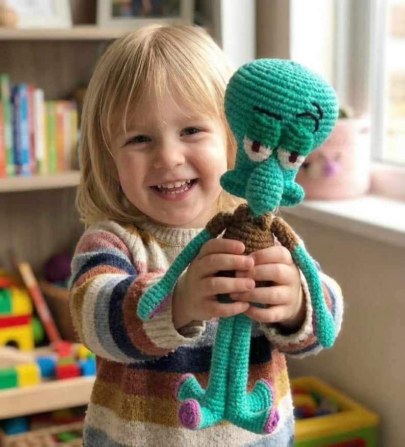 May include: A teal crochet Squidward Tentacles doll, held by a smiling child. The doll has brown and pink details. The child wears a striped jumper. The doll is a handmade item.