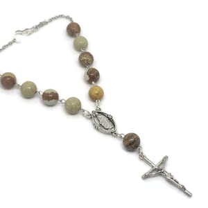 May include: A rosary featuring round, multi-colored stone beads in shades of tan, brown, and grey. The beads are connected by silver-toned metal links, leading to a central silver-toned medal depicting a figure and a silver-toned crucifix pendant.