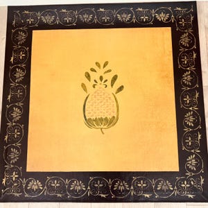 May include: A square, decorative table mat with a black border featuring a floral design. The center is a golden-yellow color with a stylized floral design in the center. The mat is likely made of a durable material.
