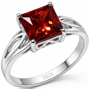 May include: A silver ring featuring a large, square-cut, red gemstone. The ring has a split shank design with decorative cutouts. The band is marked with "S925". The ring is a piece of jewelry.