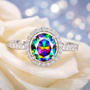 May include: A silver-toned ring featuring a large, round, multi-colored iridescent center stone. The center stone is surrounded by a halo of small, clear, sparkling stones. The band is also encrusted with small, clear, sparkling stones along the sides.