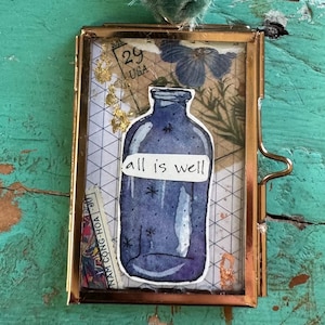 May include: A small, rectangular, gold-colored frame with a watercolor illustration of a blue bottle and the words "all is well." The frame is attached to a teal ribbon. The background features a collage of paper and botanical elements.