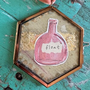 Puede incluir: Un marco hexagonal de color cobre con una ilustración de botella rosa y la palabra "float". El marco cuelga de una cinta rosa y difusa. El fondo presenta detalles de pan de oro.
