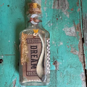 Puede incluir: Una botella de vidrio transparente con tapón de corcho, adornada con pan de oro y alambre. En el interior, una ilustración de cisne y la palabra "DREAM" son visibles en un billete vintage. La botella tiene una pequeña corona y un detalle de luna creciente.