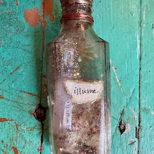 May include: A clear glass bottle with a copper-colored cap and wire detailing. The bottle contains glitter and a label that reads "illume". The background is a distressed turquoise wooden surface.