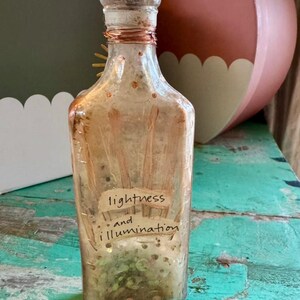 May include: A clear glass bottle with a cork stopper and copper wire detailing. The bottle contains a light-colored liquid and small particles. The label reads "lightness and illumination."