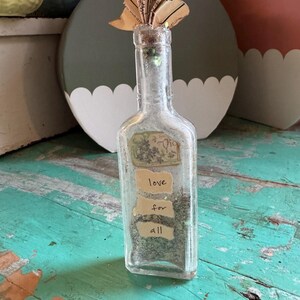 Puede incluir: Una botella de vidrio transparente con un tapón de corcho y un abanico de papel decorativo en la parte superior. La botella contiene una sustancia oscura y tiene etiquetas con las palabras "love for all". La botella está sobre una superficie de madera turquesa.
