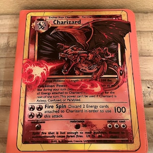 May include: A rectangular binder with a coral-colored border featuring a large graphic of the Charizard Pokémon card on the front cover. The artwork depicts a red dragon-like creature breathing fire, with visible text including Charizard, Fire Spin, and 100.