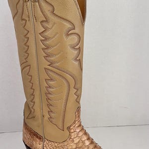 May include: Tan cowboy boot with a snakeskin foot and a leather upper. The boot features decorative stitching and a pointed toe. The heel is dark brown. A classic Western style boot.