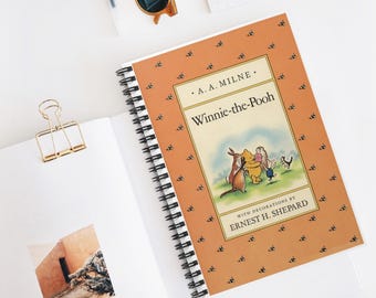 May include: A spiral-bound notebook with the cover of "Winnie-the-Pooh" by A.A. Milne, illustrated by Ernest H. Shepard. The cover is orange with a pattern of small black bees. The notebook is open on a white surface.