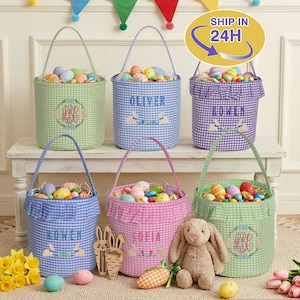 Op de afbeelding: Zes gingham paasmanden in verschillende kleuren, gevuld met kleurrijke eieren. Elke mand heeft een handvat en is gepersonaliseerd met een naam of monogram. Een pluchen konijn en houten decoraties zijn ook aanwezig.