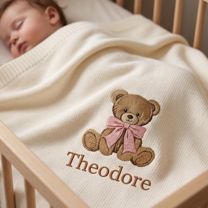 Puede incluir: Una manta de punto para bebé de color crema con un oso de peluche marrón bordado que lleva un lazo rosa en el cuello. El nombre Theodore está bordado con hilo marrón debajo del diseño del oso sobre la superficie de tela suave y texturizada.