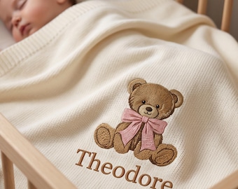 Può includere: Una coperta per neonati in maglia color crema con un orsacchiotto marrone ricamato che indossa un fiocco rosa al collo. Il nome Theodore è ricamato in filo marrone sotto il disegno dell'orsacchiotto sulla superficie in tessuto morbido e testurizzato.