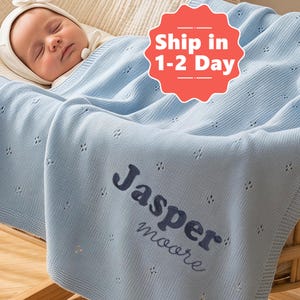 Puede incluir: Un bebé duerme plácidamente bajo una manta de punto azul claro. La manta está personalizada con el nombre "Jasper Moore" en escritura azul marino. La manta tiene un delicado diseño calado.