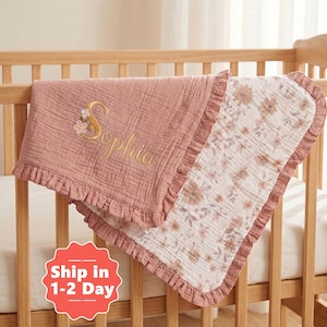 May include: A dusty rose muslin baby blanket with a ruffled edge, featuring the embroidered name Sophia in gold thread with small floral accents. The reverse side of the blanket displays a white fabric with a brown and tan floral print pattern.