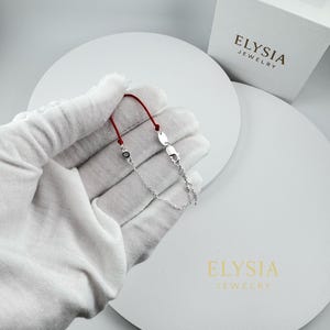May include: A silver bracelet with a red cord and a small, oval-shaped charm. The bracelet is held in a gloved hand, with a white box and the words "ELYSIA JEWELRY" visible in the background.