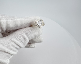 May include: A silver ring with a heart-shaped, clear gemstone center, surrounded by smaller, clear stones. The ring is held by a gloved hand against a white background. The ring is from Elysia Jewelry.