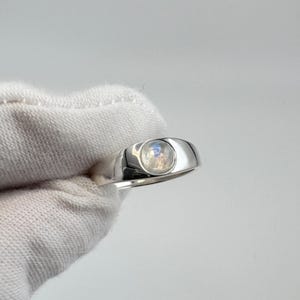 May include: A silver ring with a round, iridescent moonstone. The ring has a smooth, polished band and a bezel setting that holds the moonstone securely. The moonstone displays a soft, glowing effect, reflecting light.