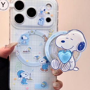 Puede incluir: Funda transparente para teléfono con un diseño de Snoopy en azul claro. La funda presenta a Snoopy en varias poses, estrellas y el texto "Happiness is a warm puppy". Una figura 3D de Snoopy con un corazón azul está adjunta.