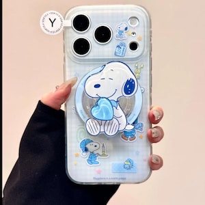May include: Clear phone case featuring a cartoon Snoopy design. The case has a light blue background with Snoopy holding a blue heart. Additional Snoopy illustrations and the text "Happiness is a warm puppy" are also visible.