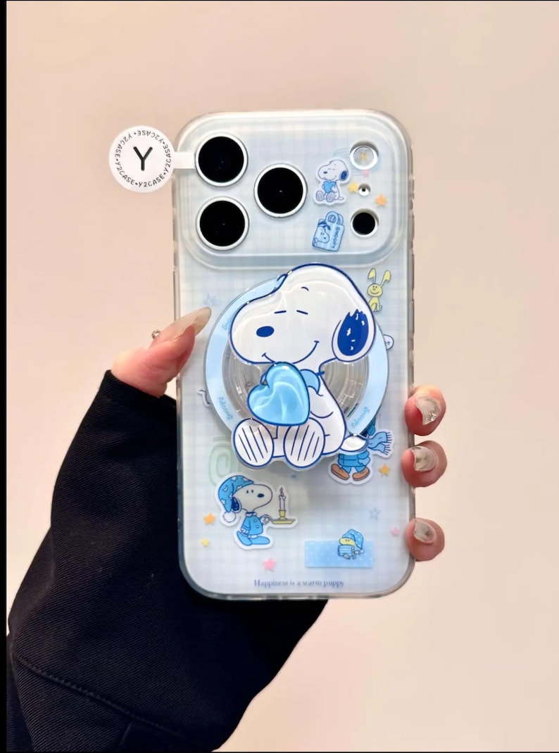 May include: Clear phone case featuring a cartoon Snoopy design. The case has a light blue background with Snoopy holding a blue heart. Additional Snoopy illustrations and the text "Happiness is a warm puppy" are also visible.