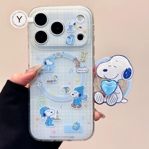 May include: Clear phone case with a Snoopy design. The case features Snoopy in various winter outfits, along with other winter-themed graphics. A matching Snoopy phone grip is attached. The case has a light blue background with the text "Happiness is a warm puppy."