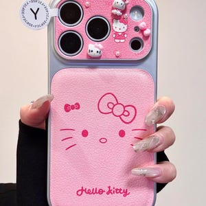 May include: Pink phone case featuring a Hello Kitty design. The case has a silver frame and includes 3D Hello Kitty figures and the text "Hello Kitty". The case is designed for a smartphone.