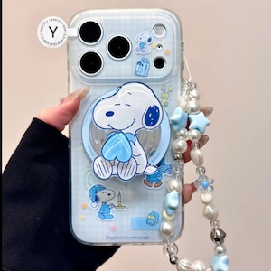May include: Clear phone case featuring a Snoopy design with blue accents. The case has a blue grid pattern and includes a beaded wrist strap with blue star and pearl accents. The text "Happiness is a warm puppy" is visible.