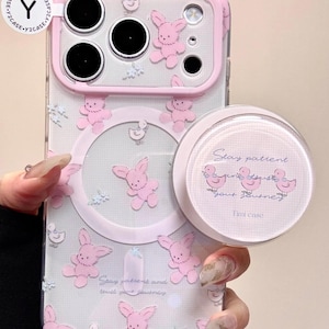 Puede incluir: Funda de teléfono transparente con borde rosa, con diseños de conejitos y patos rosas. Un pop-socket redondo a juego con el texto "Stay patient your journey" está adjunto. La funda tiene una estética linda y caprichosa.