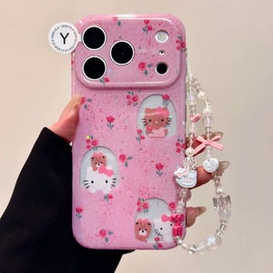 Pu&ograve; includere: Custodia per telefono rosa con motivo floreale e disegni di Hello Kitty. La custodia ha una finitura glitterata e un cinturino da polso con perline con ciondoli, tra cui fiocchi e un ciondolo "Good Luck".