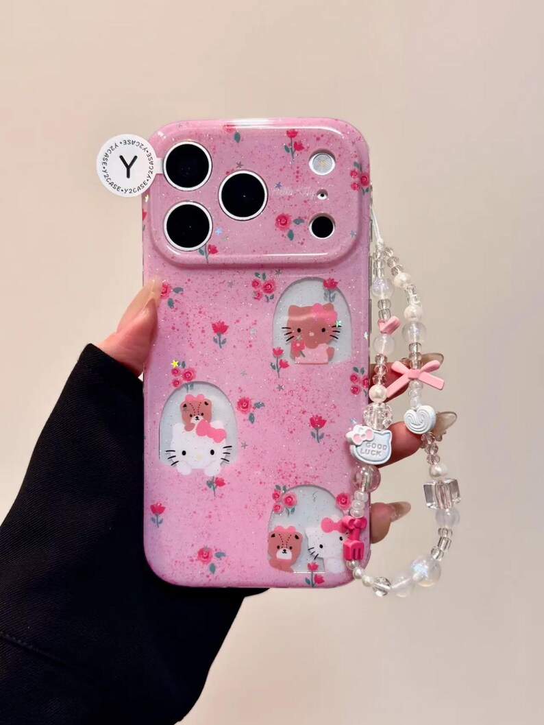 Pu&ograve; includere: Custodia per telefono rosa con motivo floreale e disegni di Hello Kitty. La custodia ha una finitura glitterata e un cinturino da polso con perline con ciondoli, tra cui fiocchi e un ciondolo "Good Luck".