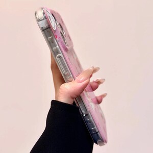 Pu&ograve; includere: Una custodia trasparente per telefono con un design floreale rosa e piccoli cuori. La custodia ha un bordo trasparente ed &egrave; tenuta in diagonale, mostrando il design e i pulsanti laterali del telefono.