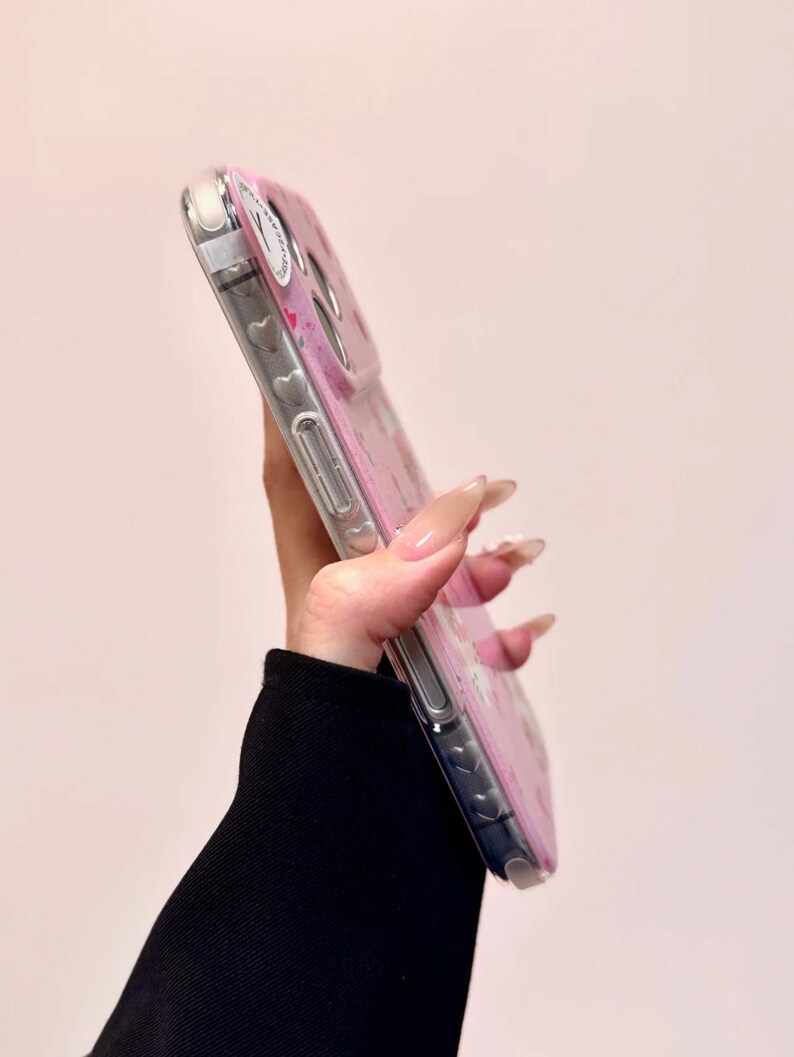 Pu&ograve; includere: Una custodia trasparente per telefono con un design floreale rosa e piccoli cuori. La custodia ha un bordo trasparente ed &egrave; tenuta in diagonale, mostrando il design e i pulsanti laterali del telefono.