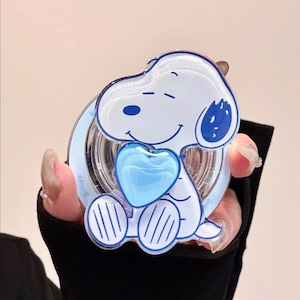 May include: A Snoopy-themed compact mirror. The mirror features a white Snoopy design with blue accents, holding a blue heart. The mirror has a clear, circular frame with the word "Snoopy" printed on it. The overall design is playful and cartoonish.
