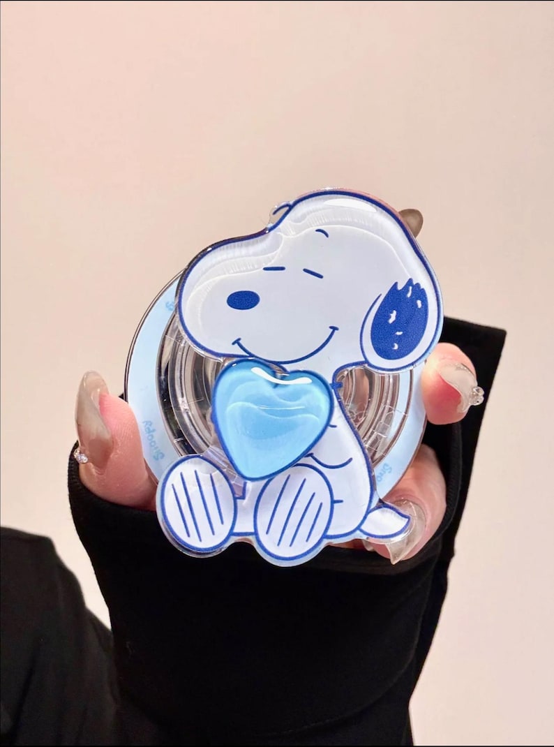 May include: A Snoopy-themed compact mirror. The mirror features a white Snoopy design with blue accents, holding a blue heart. The mirror has a clear, circular frame with the word "Snoopy" printed on it. The overall design is playful and cartoonish.