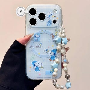 May include: Clear phone case with a blue and white Snoopy design. The case features Snoopy illustrations, stars, and the text "Happiness is a warm puppy." A beaded strap with blue stars and pearls is attached.