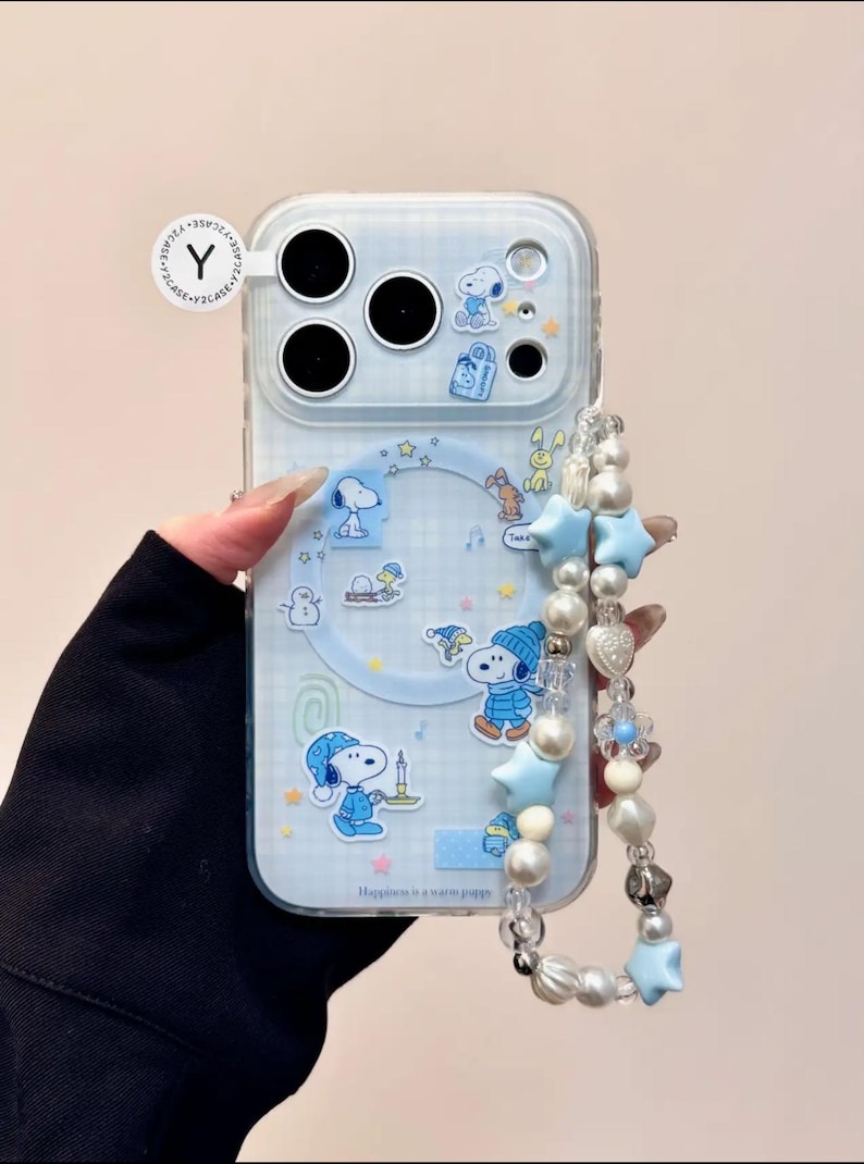May include: Clear phone case with a blue and white Snoopy design. The case features Snoopy illustrations, stars, and the text "Happiness is a warm puppy." A beaded strap with blue stars and pearls is attached.