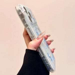 May include: Clear phone case with a light blue design featuring cartoon characters. The case has a transparent border and a glossy finish. The design includes heart-shaped details along the side.