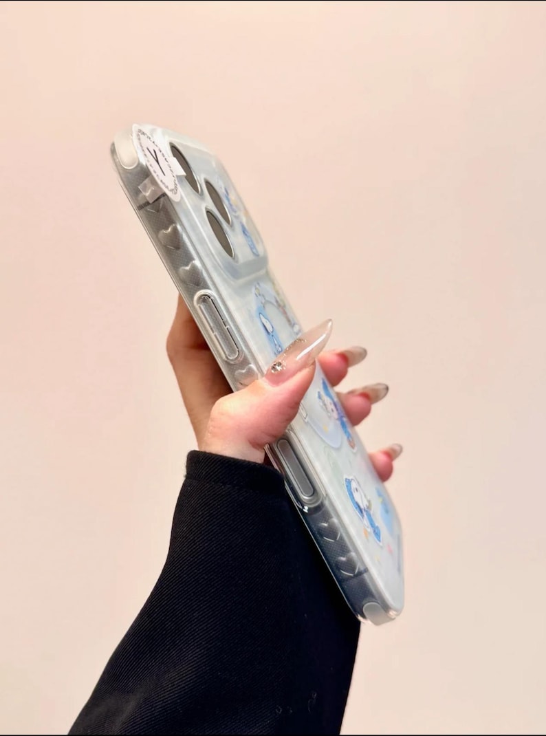 May include: Clear phone case with a light blue design featuring cartoon characters. The case has a transparent border and a glossy finish. The design includes heart-shaped details along the side.