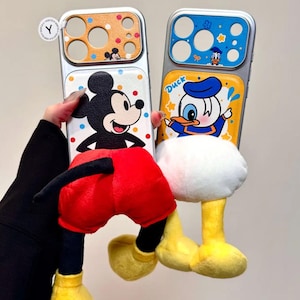 May include: Two phone cases featuring Disney characters. One case has a Mickey Mouse design with red shorts and yellow shoes. The other case has a Donald Duck design with a blue hat and yellow feet. Both cases have a silver frame.