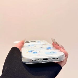 May include: Clear phone case with a cartoon character design in blue. The case has a transparent border and features a series of small holes and openings for the phone's features. The design includes various cartoon figures and shapes.