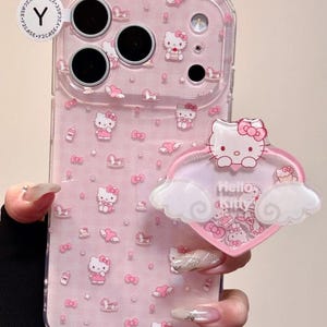 Puede incluir: Funda de teléfono rosa con diseño de Hello Kitty. La funda presenta un patrón repetido de personajes de Hello Kitty, lazos y corazones. Un soporte en forma de corazón con Hello Kitty y la inscripción "Hello Kitty" está adjunto.