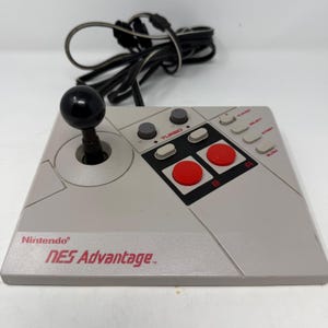 Puede incluir: Un mando de estilo arcade Nintendo NES Advantage de color gris con un joystick negro, dos botones rojos grandes A y B, dos diales turbo grises y botones grises adicionales etiquetados como Player, Select, Start y Slow, con un cable negro.