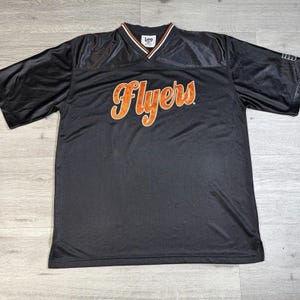 May include: Black jersey with the word "Flyers" in orange script across the front. The jersey has a V-neck with white and orange trim. The sleeves are short and the material appears to be a mesh fabric.