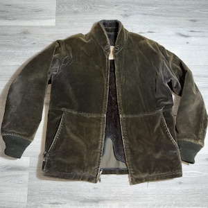 Op de afbeelding: Een donker olijfgroene corduroy jas met een hoge kraag en een ritssluiting over de hele lengte. De jas heeft twee voorzakken en geribde manchetten. De binnenvoering is donkerder bruin. De jas is een medium maat.