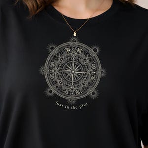 May include: Black t-shirt featuring a detailed, circular compass design in white. The design includes a compass rose and intricate patterns, with the text "lost in the plot" below. The shirt is a classic crew neck style.