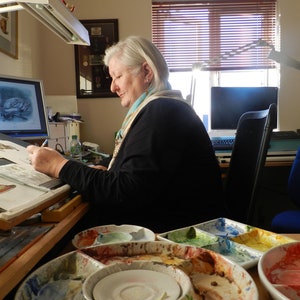 Peut inclure: Une femme est assise &agrave; un bureau avec un &eacute;cran d'ordinateur montrant un dessin de cygne. Elle tient un crayon et travaille sur un dessin. Il y a plusieurs palettes de peinture avec de la peinture s&eacute;ch&eacute;e dessus.