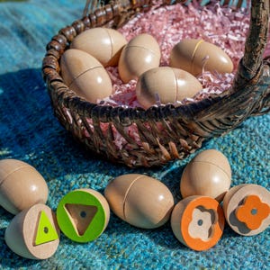 May include: A wicker basket filled with wooden eggs and pink paper shreds. Several wooden eggs are scattered around the basket. Some of the eggs have colorful geometric shapes on their ends, including a triangle and a flower.